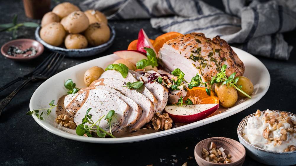 Kalkunbryst med grønnsaker og poteter på en hvit tallerken, omgitt av små boller med saus og krydder, på et mørkt bord.