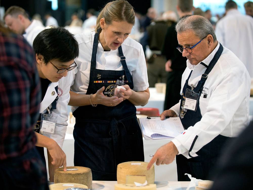 Dommere vurderer ost under oste-VM 2023, med flere personer i hvite frakker og en person med en notatblokk.