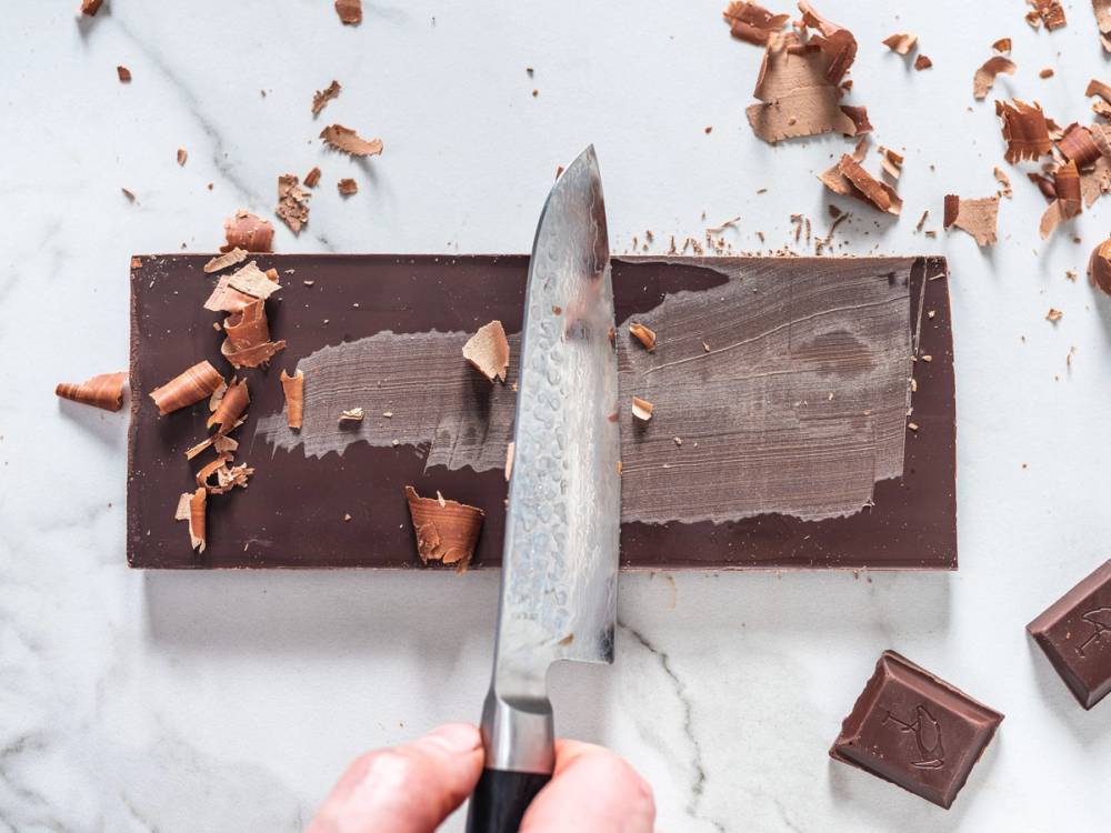 En person skjærer et stykke mørk sjokolade med en kniv på et hvitt underlag, med sjokoladekrummel rundt.