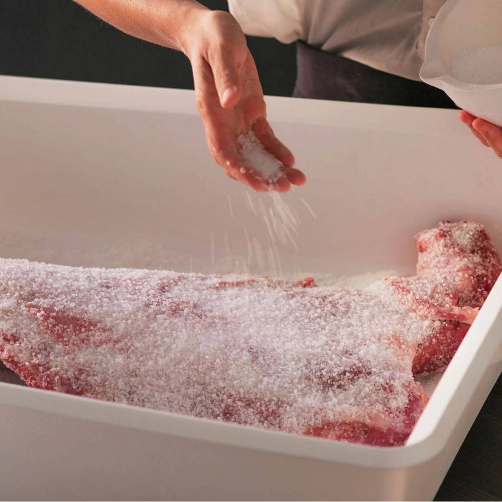 Person som drys salt over et stykke pinnekjøtt i en stor hvit plastkasse, klar til å tilberedes.