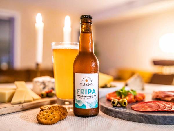 En flaske alkoholfri juleøl står på et bord sammen med et glass med øl, omgitt av julemat og kjente juledekorasjoner i bakgrunnen.