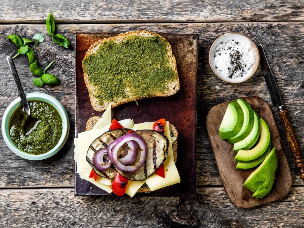 En skive med pesto, en skive brød med ost, skiver av agurk og en liten bolle med smør eller krem, alt plassert på en treplate på et rustikt trebord.