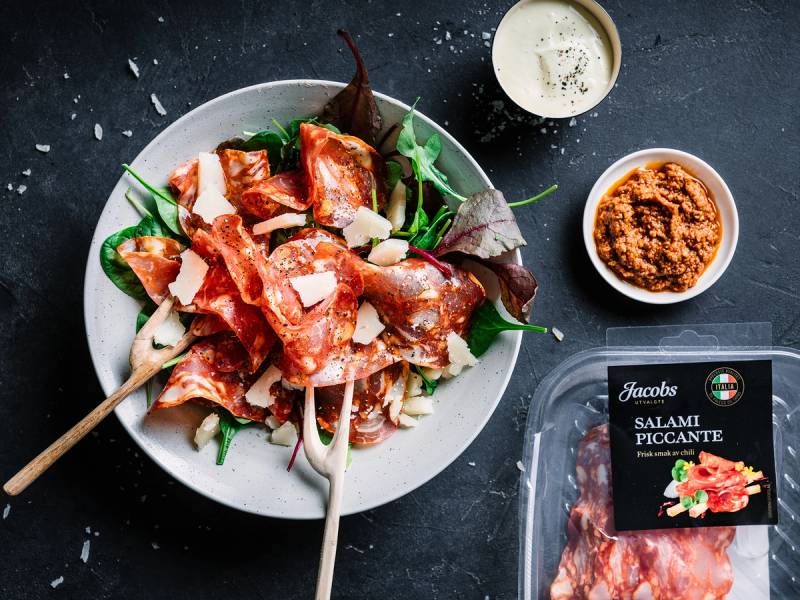 Skål med spekemat og salami, samt små boller med krydder og saus på et mørkt bord