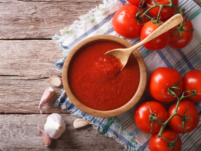 Tomatsaus i en bolle med friske tomater og hvitløk rundt på et trebord.