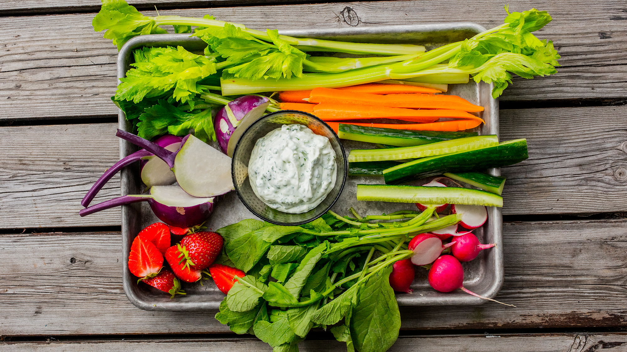 Et utvalg av friske grønnsaker som gulrøtter, reddiker, agurk, tomater, og ulike bladgrønnsaker på et trebrett.