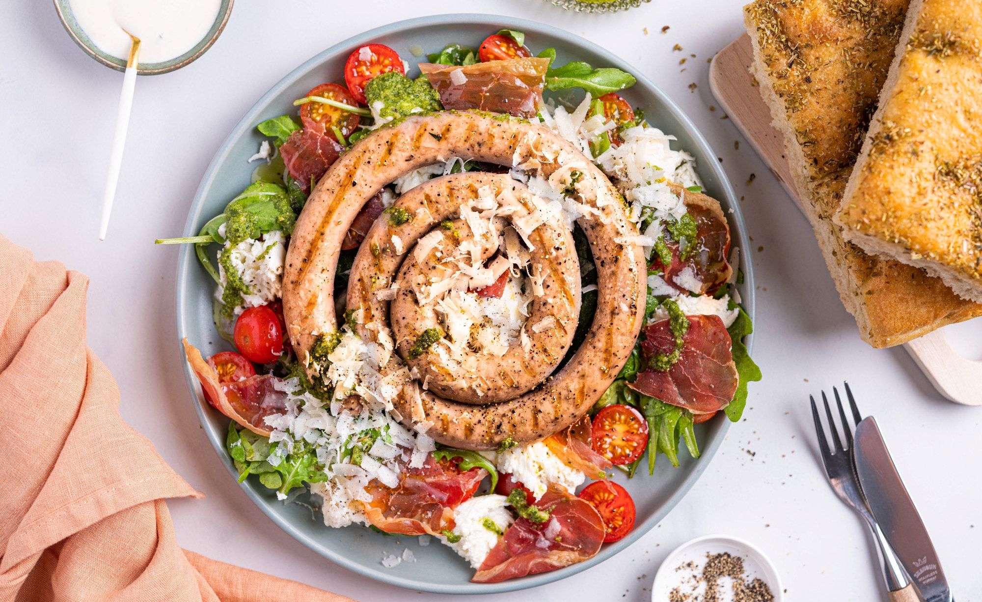 Salsiccia salat med mozzarella og parmaskinke på en tallerken med cherrytomater og grønt, omgitt av brød og bestikk.