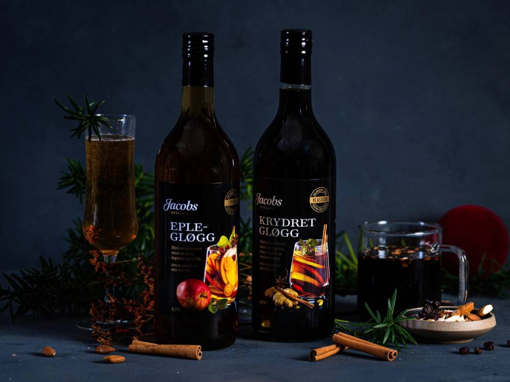 To flasker med Jacobs gløgg, en glass med gløgg, og krydder som kanelstenger og nellik rundt på et mørkt bord med juledekorasjoner.