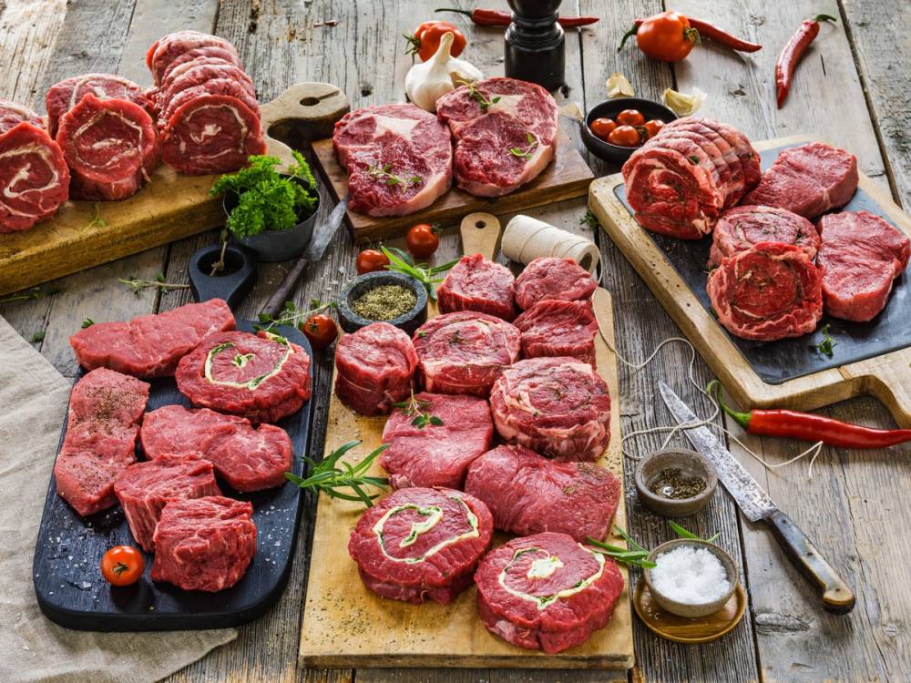 Ulike typer av rått kjøtt, inkludert biffer og steker, lagt ut på tre skjærebrett og serveringsfat med krydder og urter på en rustikk treflate.