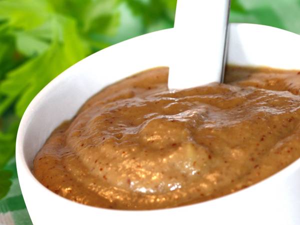 Gyllen sennep i en hvit skål med en skje oppi, mot en grønn bakgrunn.