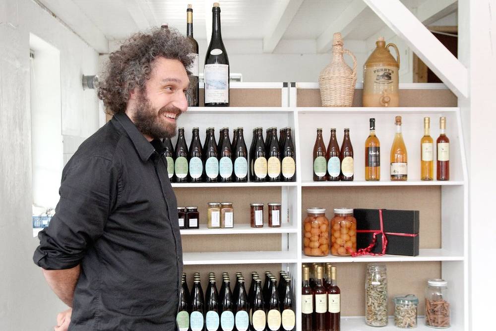 En person står i en butikk med hyller fylt med ulike flasker og glass, muligens en vin- eller delikatessebutikk.