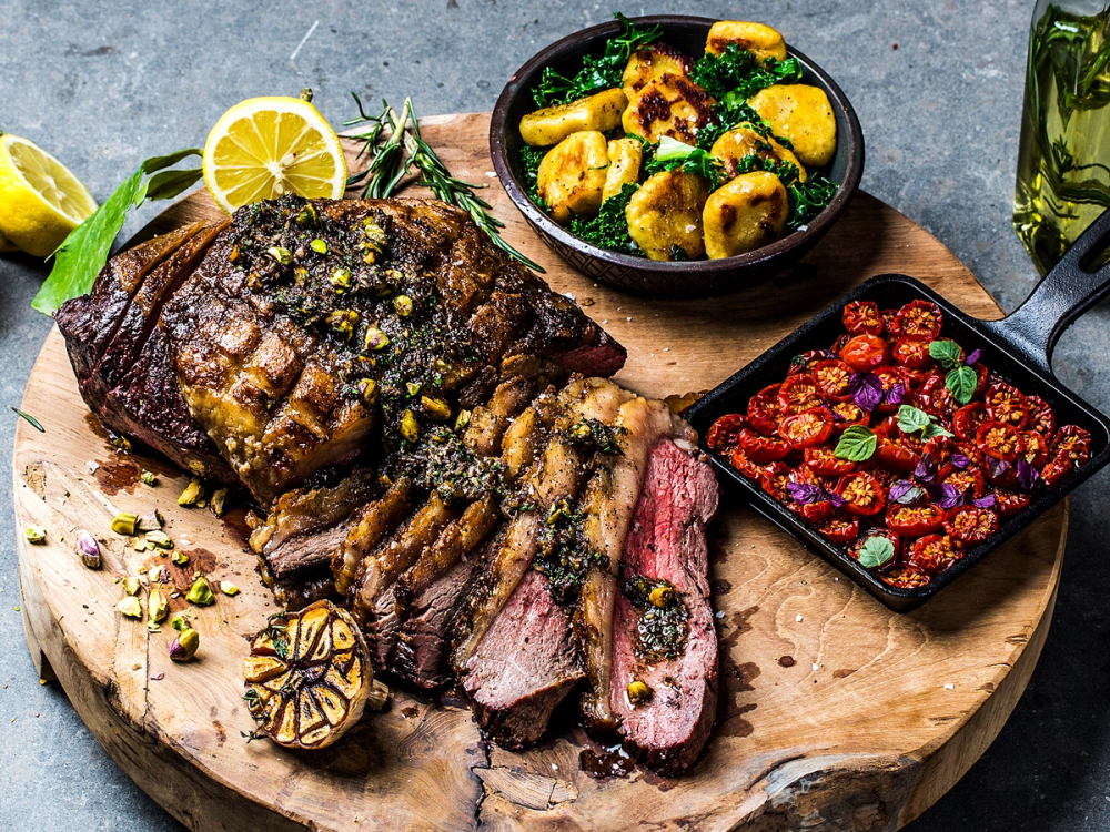 En skive av stekt biff med rosa midt, servert med en sitronbåt og krydder på en treplate, sammen med en grønnsakssalat og en liten gryte med bærkompott.