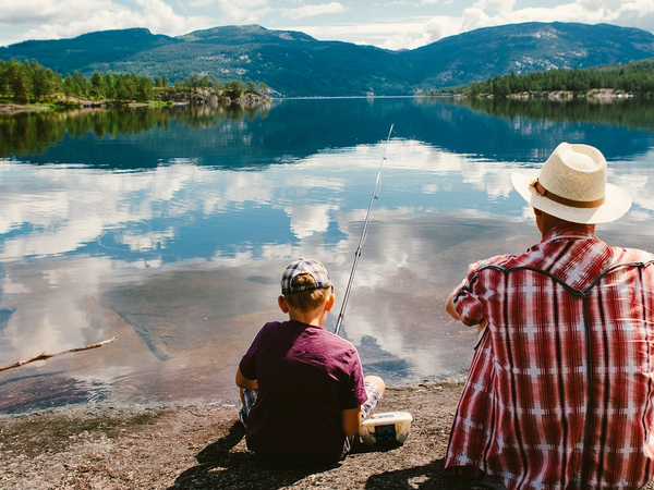 En voksen og et barn sitter ved vannkanten og ser ut over en innsjø med fjell i bakgrunnen.