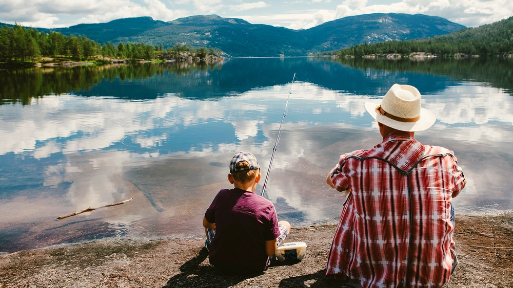 En voksen og et barn sitter ved vannkanten og ser ut over en innsjø med fjell i bakgrunnen.