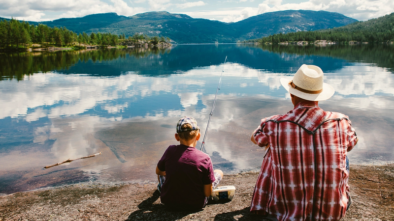 En voksen og et barn sitter ved vannkanten og ser ut over en innsjø med fjell i bakgrunnen.