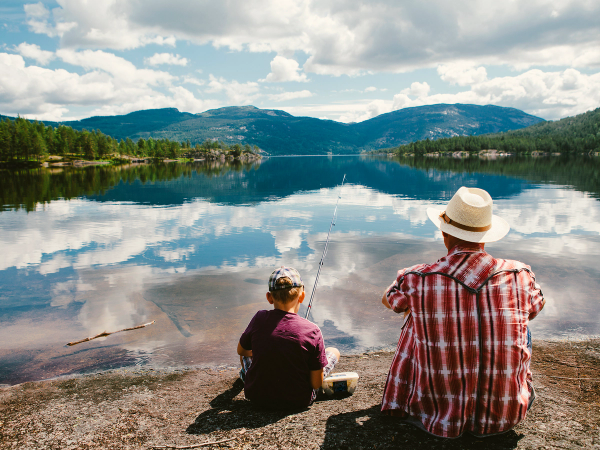 En voksen og et barn sitter ved vannkanten og ser ut over en innsjø med fjell i bakgrunnen.