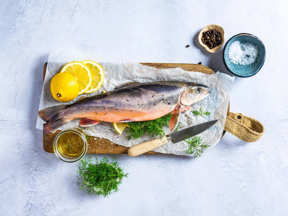 En fersk telemarkrøye ligger på et skjærebrett med sitron, urter og en liten skål med krydder ved siden av.