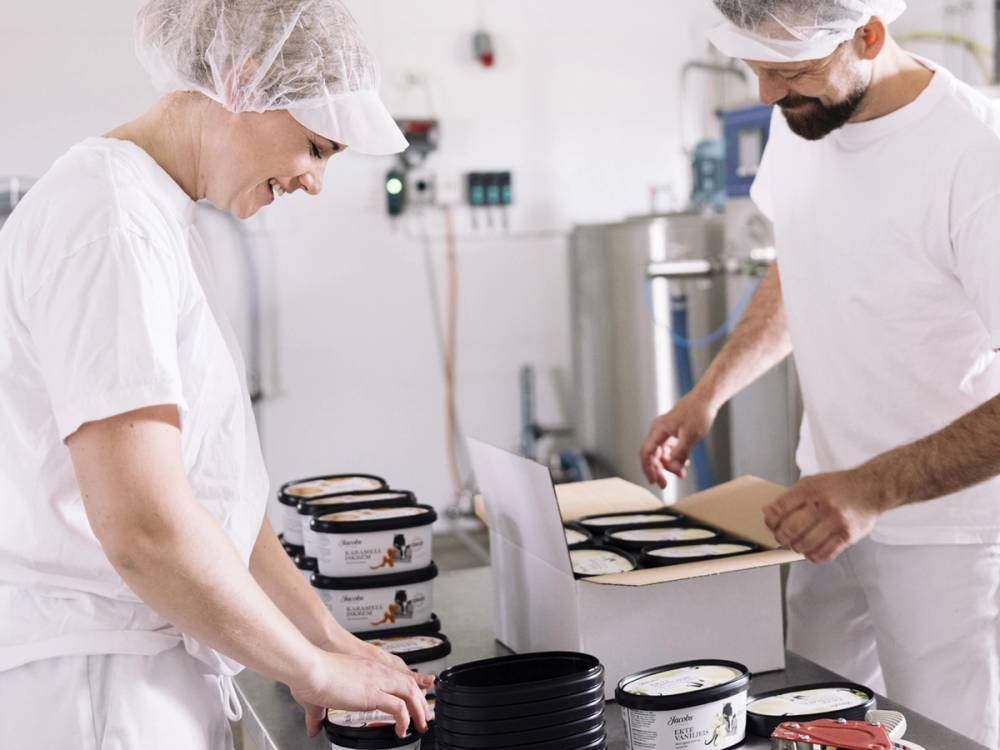 To personer pakker iskremkasser i en produksjonslinje på en iskremfabrikk, med iskremkasser og utstyr rundt seg.