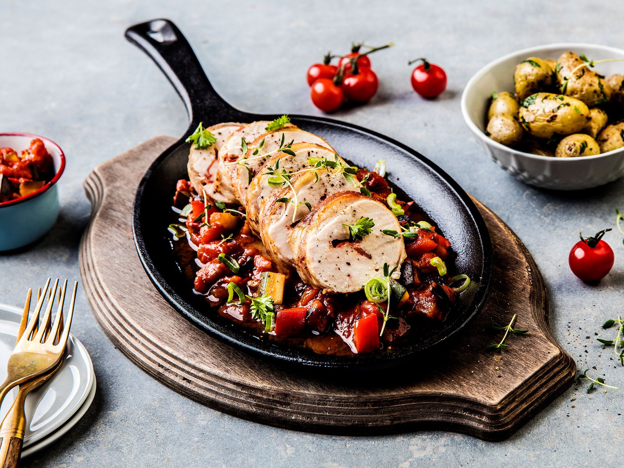 Saftig kyllingbryst med chorizo og chaumes på en svart stekepanne med cherrytomater rundt, på et rundt trefat.