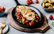 Saftig kyllingbryst med chorizo og chaumes på en svart stekepanne med cherrytomater rundt, på et rundt trefat.