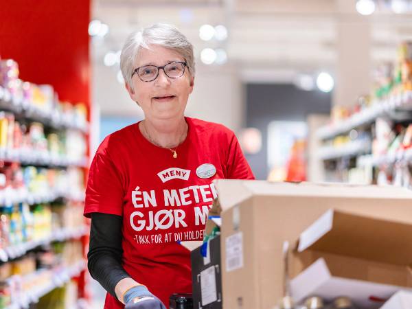 En person i rød T-skjorte som jobber i en butikk, med hyller fylt med varer i bakgrunnen.