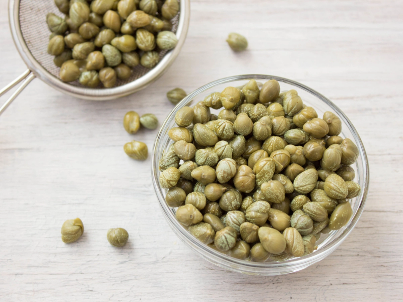 To glasskåler med grønne erter på et lyst bord, hvorav den ene er større og fylt med erter, og noen er spredt rundt.