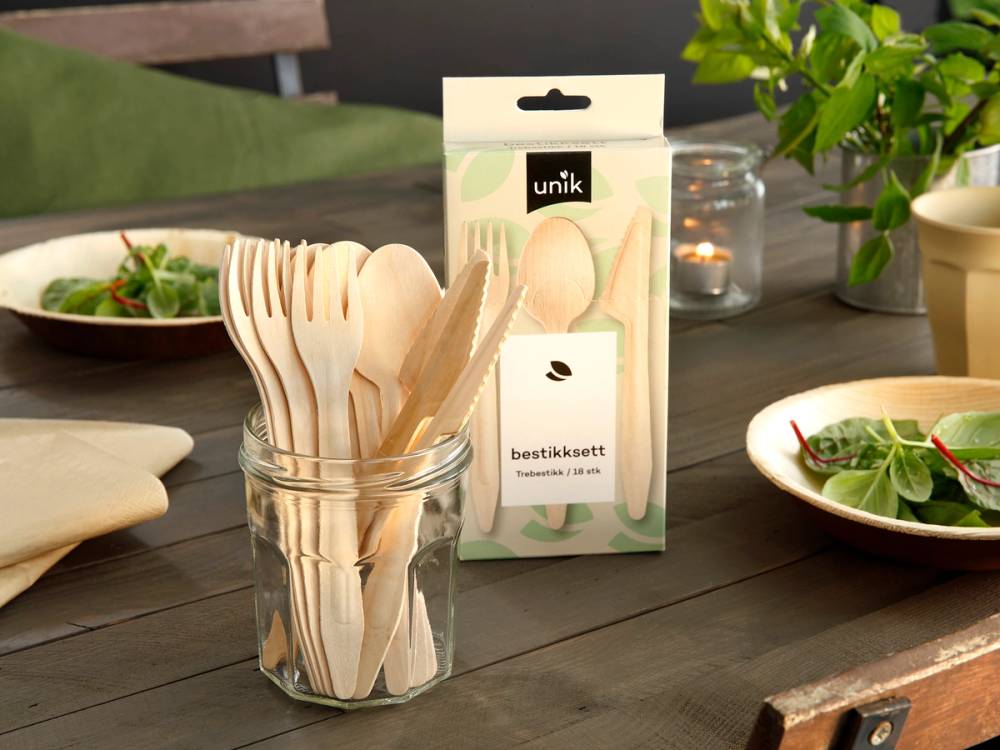 En glasskrukke med tretten trepinner og en pakke med bestikksett på et bord med grønne planter og salat i bakgrunnen.