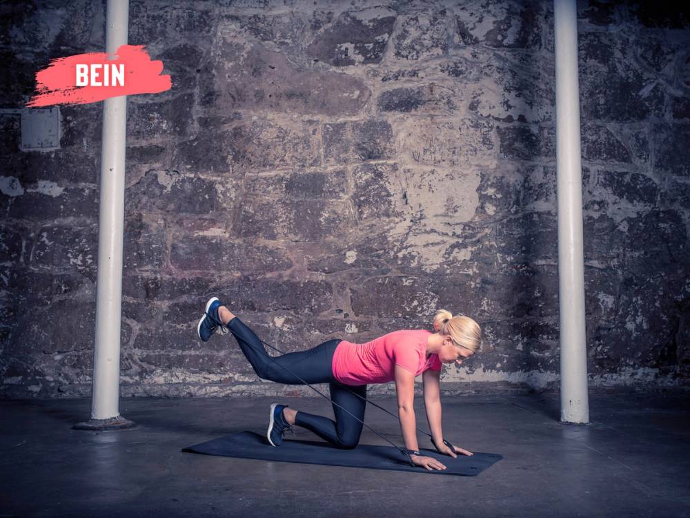 En kvinne gjør en øvelse på gulvet i et treningsrom med en murvegg i bakgrunnen, iført rosa treningstøy og svarte treningsklær.
