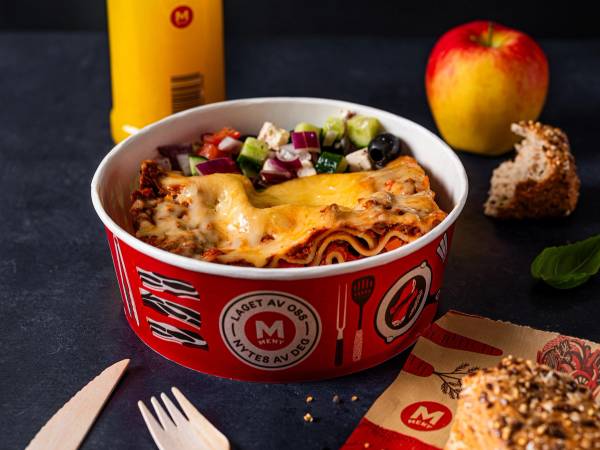 En porsjon lasagne i en rød og hvit emballasje med en gul ketchupflaske, et eple og et stykke brød ved siden av på et bord.