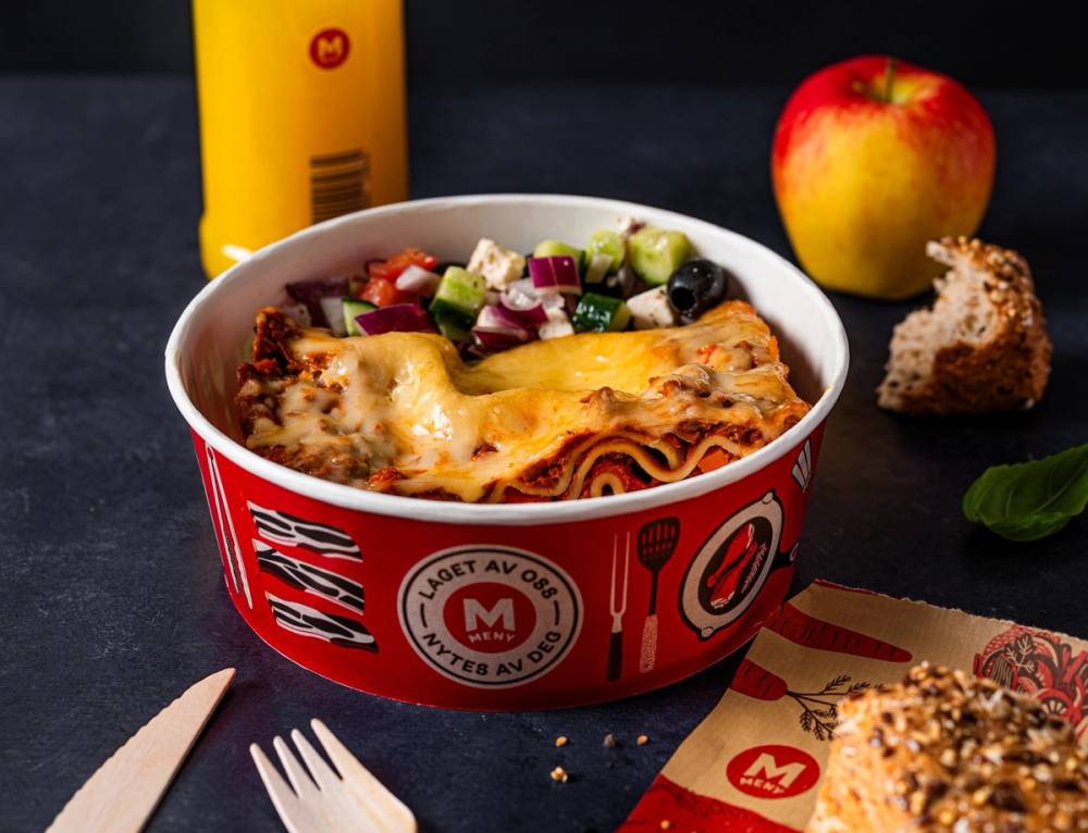 En porsjon lasagne i en rød og hvit emballasje med en gul ketchupflaske, et eple og et stykke brød ved siden av på et bord.