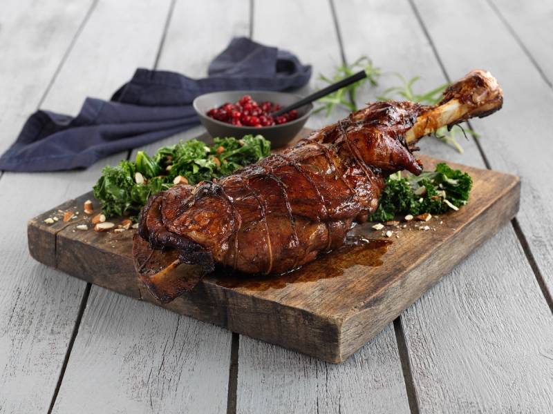 Stekt kje med urter og krydder, servert på et trebrett med grønnsaker og en liten skål med røde bær, på et lyst bord.