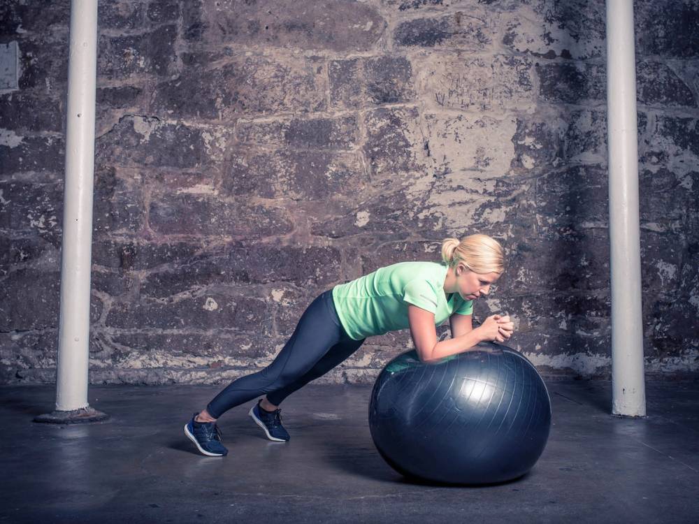 En kvinne trener med en stor treningsball i et treningsrom med murvegg og stålstolper.