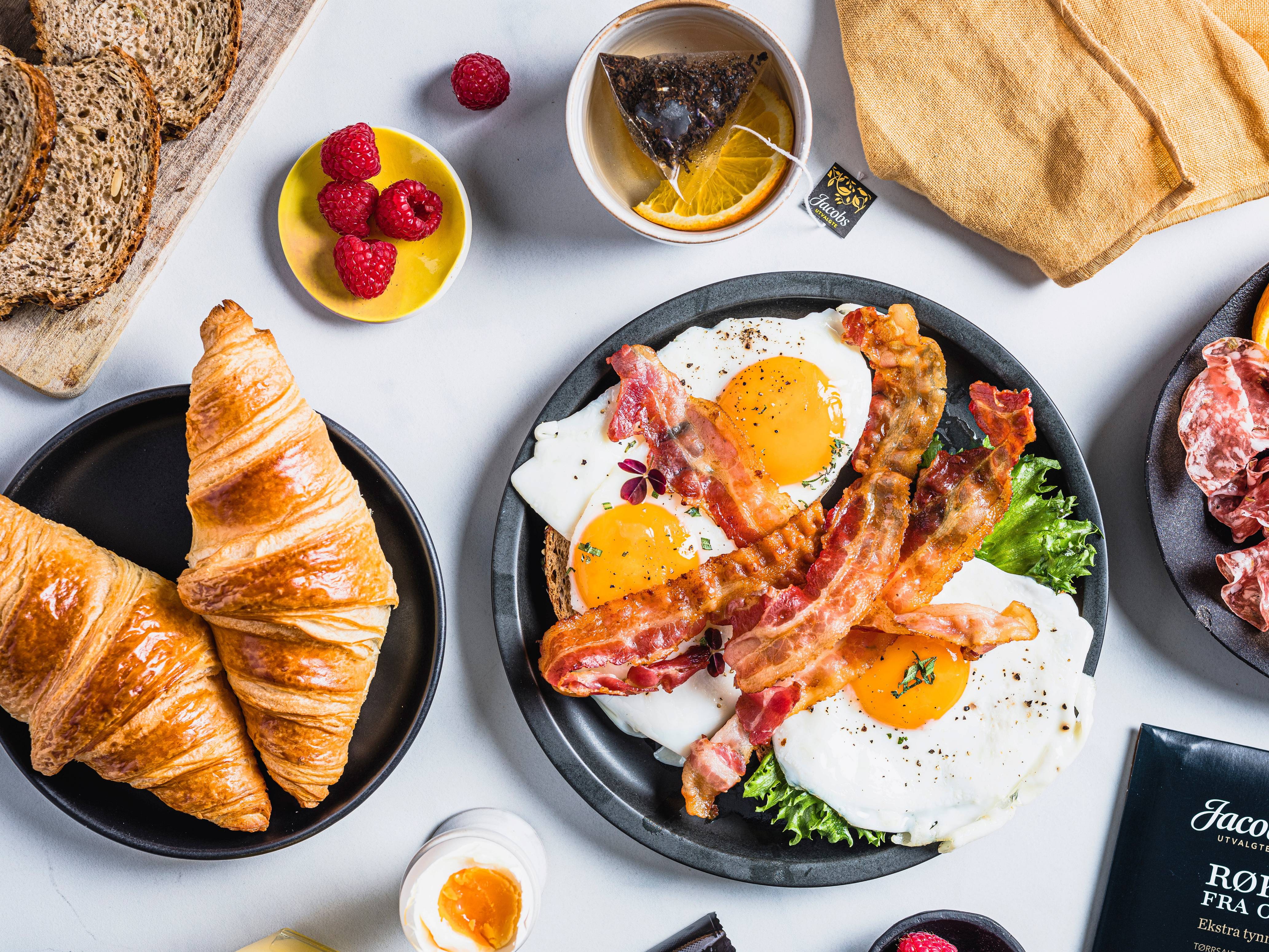 Frokost med to stekte egg, bacon, croissants, brød, og bær på en hvit bakgrunn.