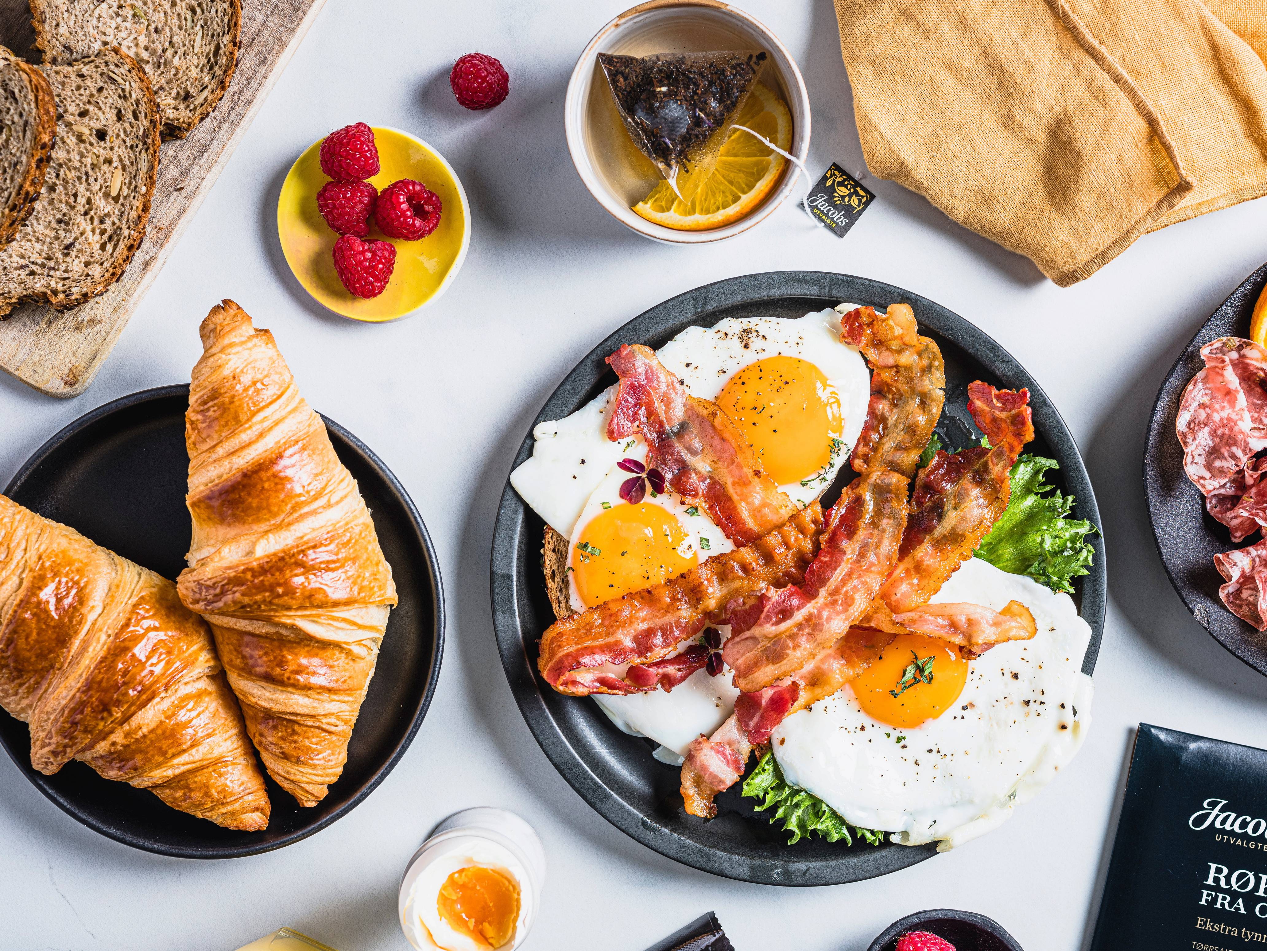 Frokost med to stekte egg, bacon, croissants, brød, og bær på en hvit bakgrunn.