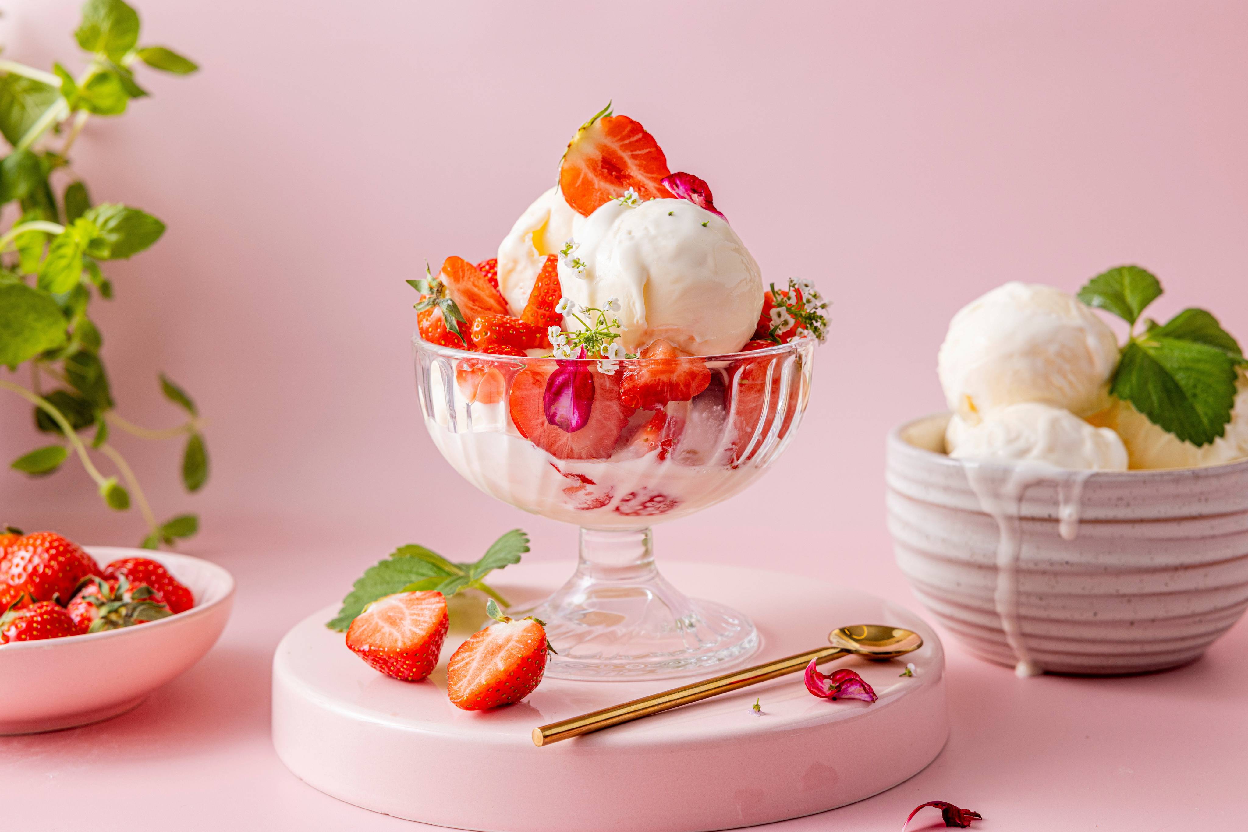 Iskrem med jordbær i en glasskål, omgitt av friske jordbær og mynte på en rosa bakgrunn.