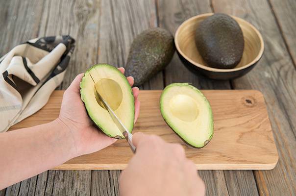 En person skreller en avokado med en skje på et skjærebrett, med to hele avokadoer i en skål i bakgrunnen.