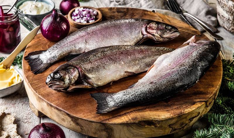 Tre hele rakfisk ligger på et rundt trebrett med små glass med tilbehør og rødløk rundt, klar til servering til jul.