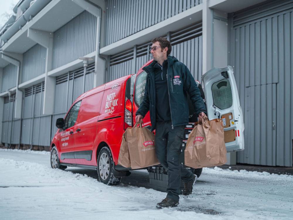 En person bærer flere handleposer ut av en varebil foran en industriell bygning, med snø på bakken.