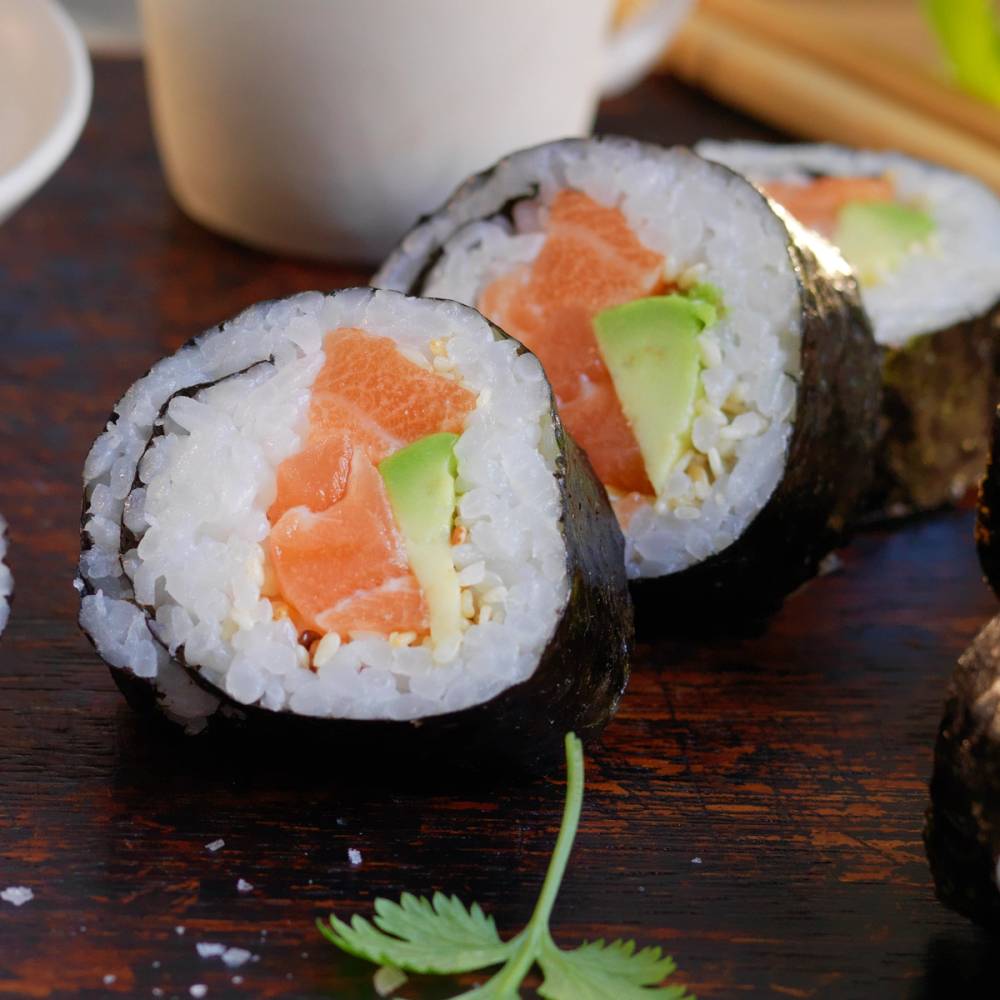To maki sushi-ruller med laks, avokado og ris på en mørk treplate, med en liten grønn urt ved siden av.
