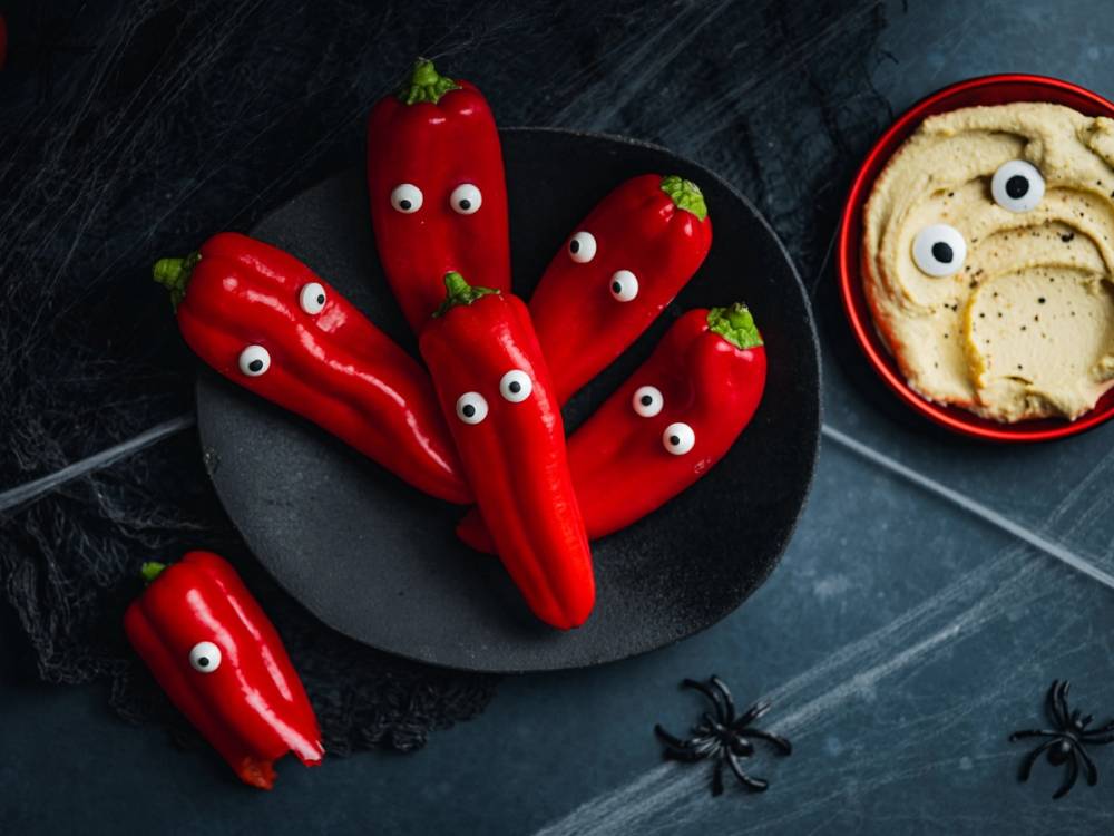 Røde paprika med googlyøyne på en svart tallerken, og en skål med kjeks med øyne i bakgrunnen.