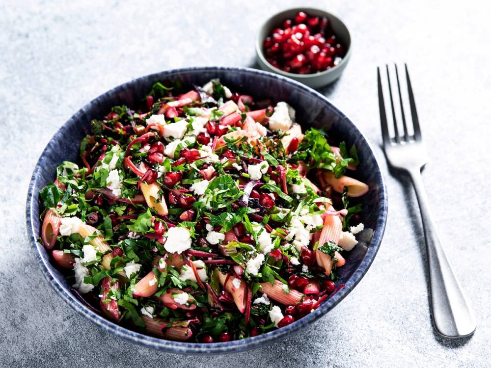 En skål med pastasalat blandet med grønnkål, granateple og nøtter, med en liten skål med granatepler ved siden av og en gaffel på tallerkenen.
