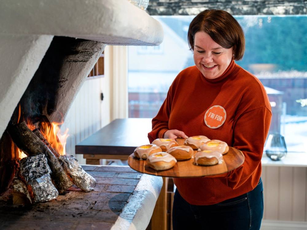 En person med kort hår som holder en tallerken med flere små kaker foran en peis med brennende ild, i et rom med store vinduer.