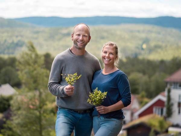 To personer står ute i naturen og holder blomster, med landskap og hus i bakgrunnen.
