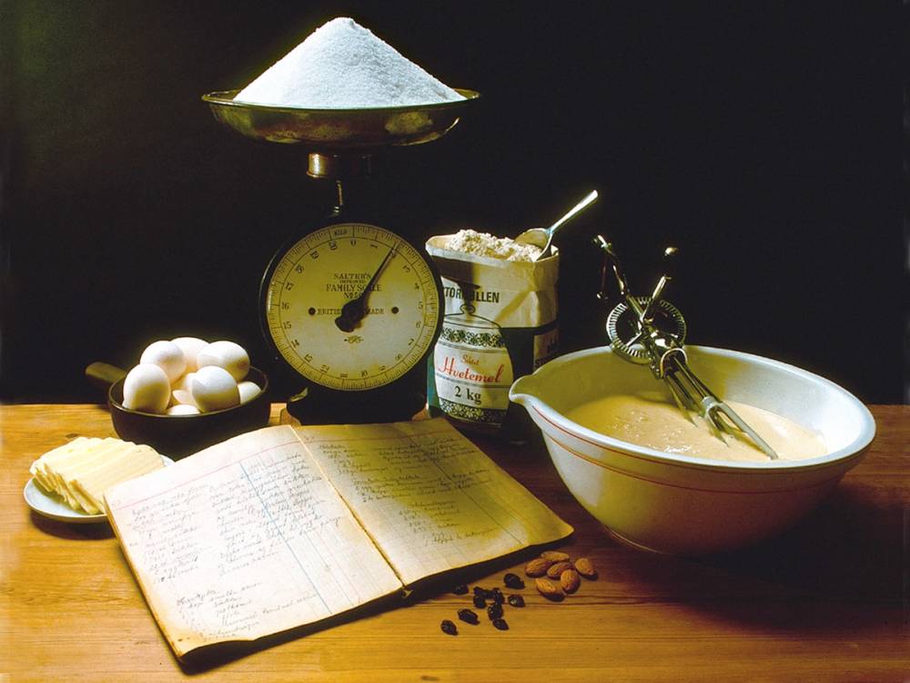 En kjøkkenvekt, en åpen kokebok, egg, kaffebønner og en bolle med kjøkkenredskaper på et bord, med en skål og en boks med ingredienser i bakgrunnen.