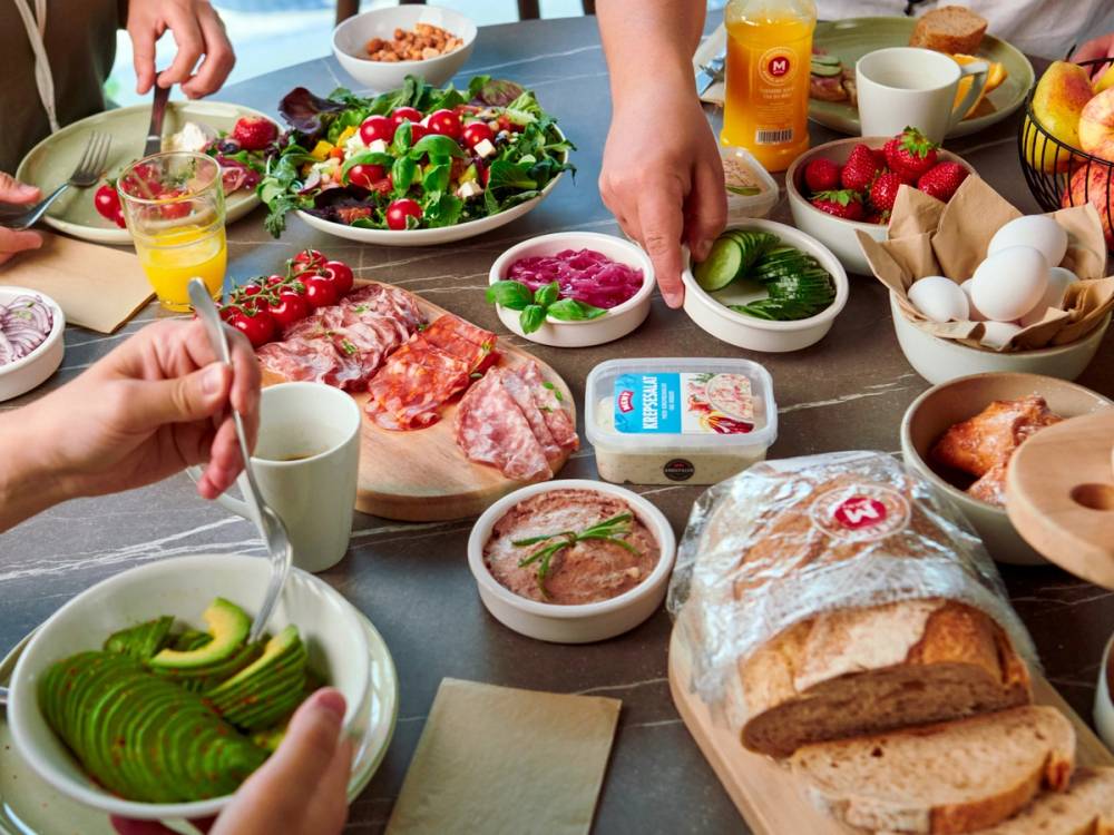 Folk som spiser frokost med brød, pålegg, frukt og juice rundt et bord.