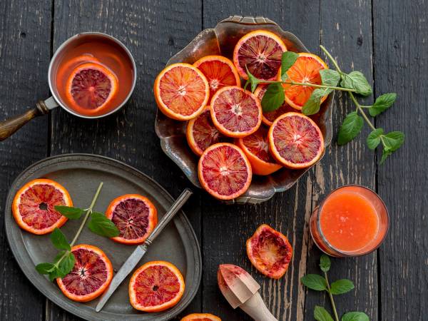 Skiver av blodappelsin med rødt kjøtt og grønne blader på en tallerken og et glass med blodappelsinjuice på en mørk treflate