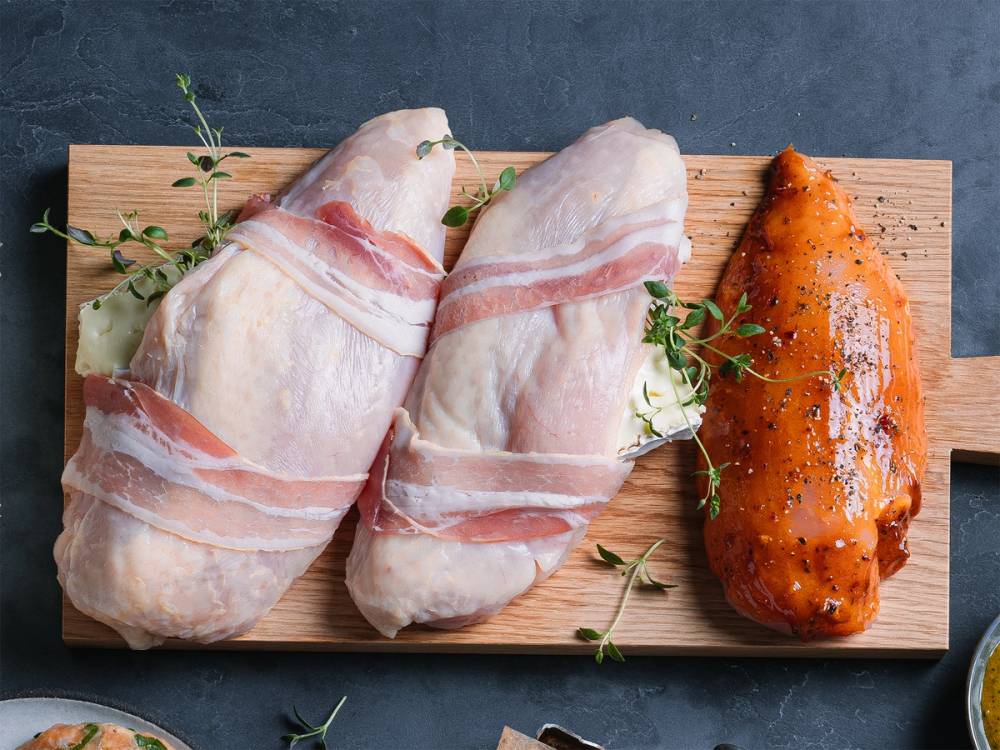 To rå kyllingfileter innpakket i bacon på et skjærebrett med urter ved siden av.