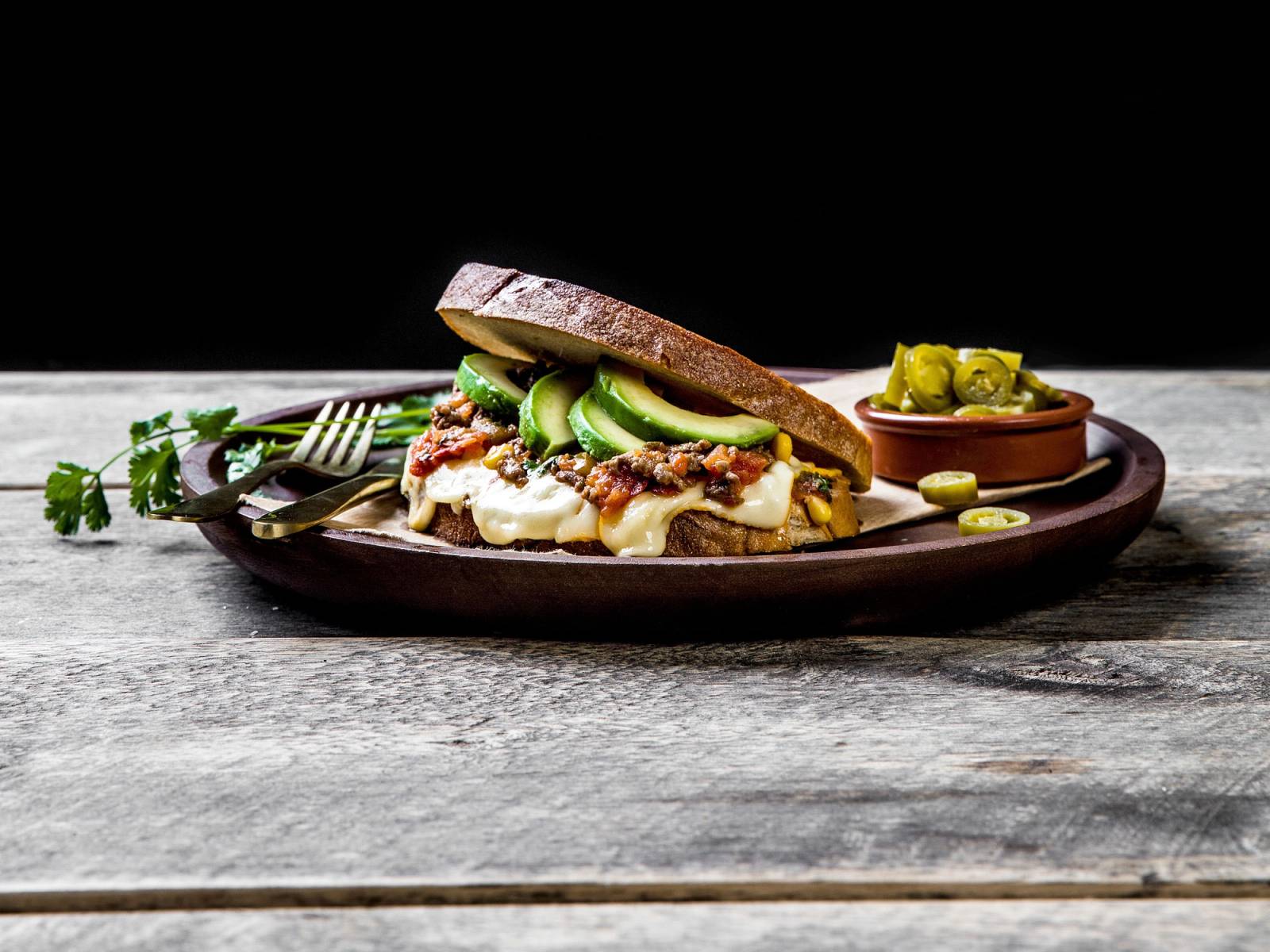 Ostesmørbrød med avokado og ost på en tallerken, med en liten skål med tilbehør ved siden av, på en treplate mot en svart bakgrunn.
