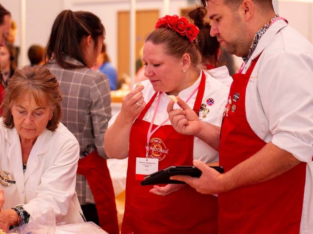 En gruppe mennesker, inkludert en person i hvit kokkebekledning og en kvinne med rød blomsterhårbånd, snakker sammen på et arrangement.