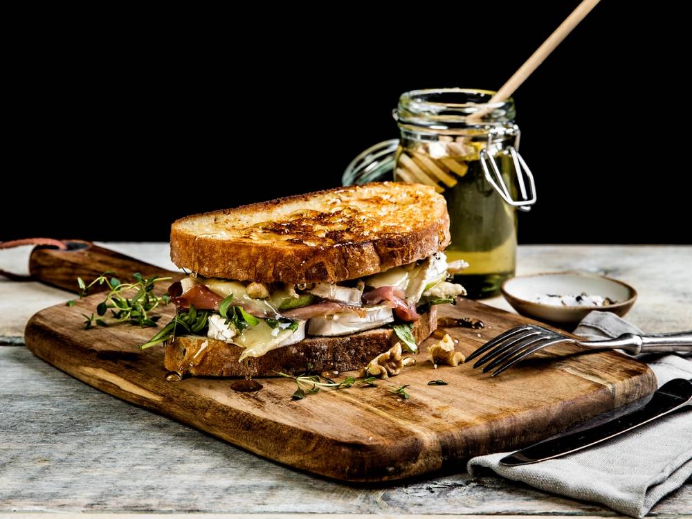 Ostesmorbrod med chevreost og urter på en skjærefjøl med en skje med honning i bakgrunnen.