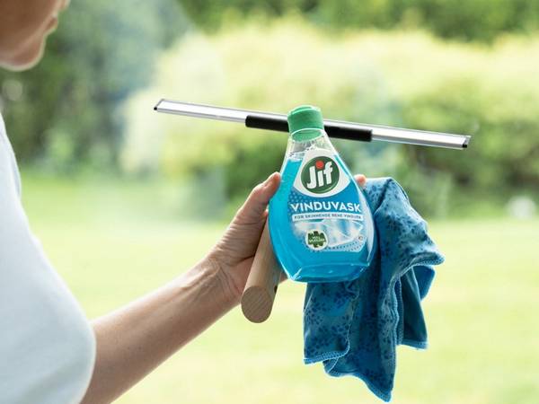 En person holder et vindusvaskeredskap med en flaske med vindusvask og en klut, foran et utendørs landskap med grønt gress og trær.